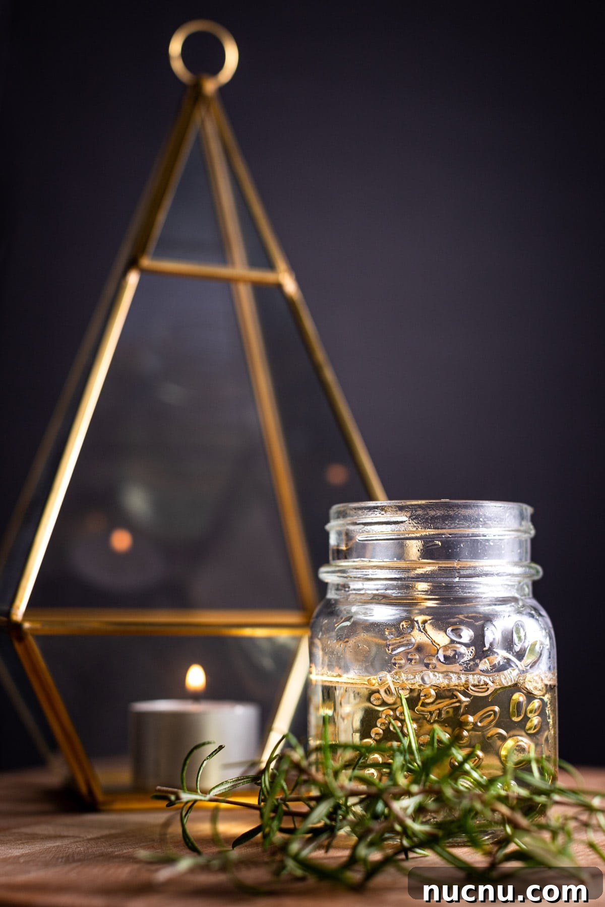 Jar of rosemary simple syrup, on a wooden board, next to a candle, gold votive and fresh rosemary on a black background.