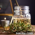 A small glass jar filled with rosemary syrup, with fresh rosemary in front and a candle in the background.