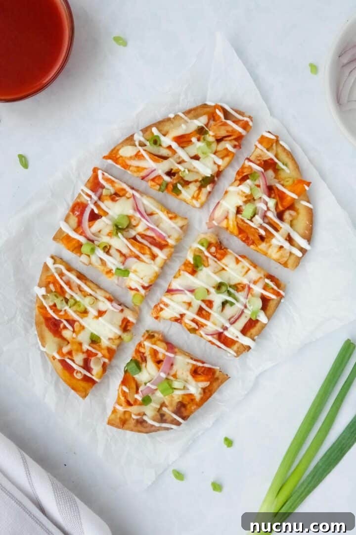 Spicy Buffalo Chicken Flatbread Pizza Recipe for Game Day 2 Whole buffalo chicken flatbread topped with green onions, red onions, and a drizzle of sour cream displayed on parchment paper on a white counter