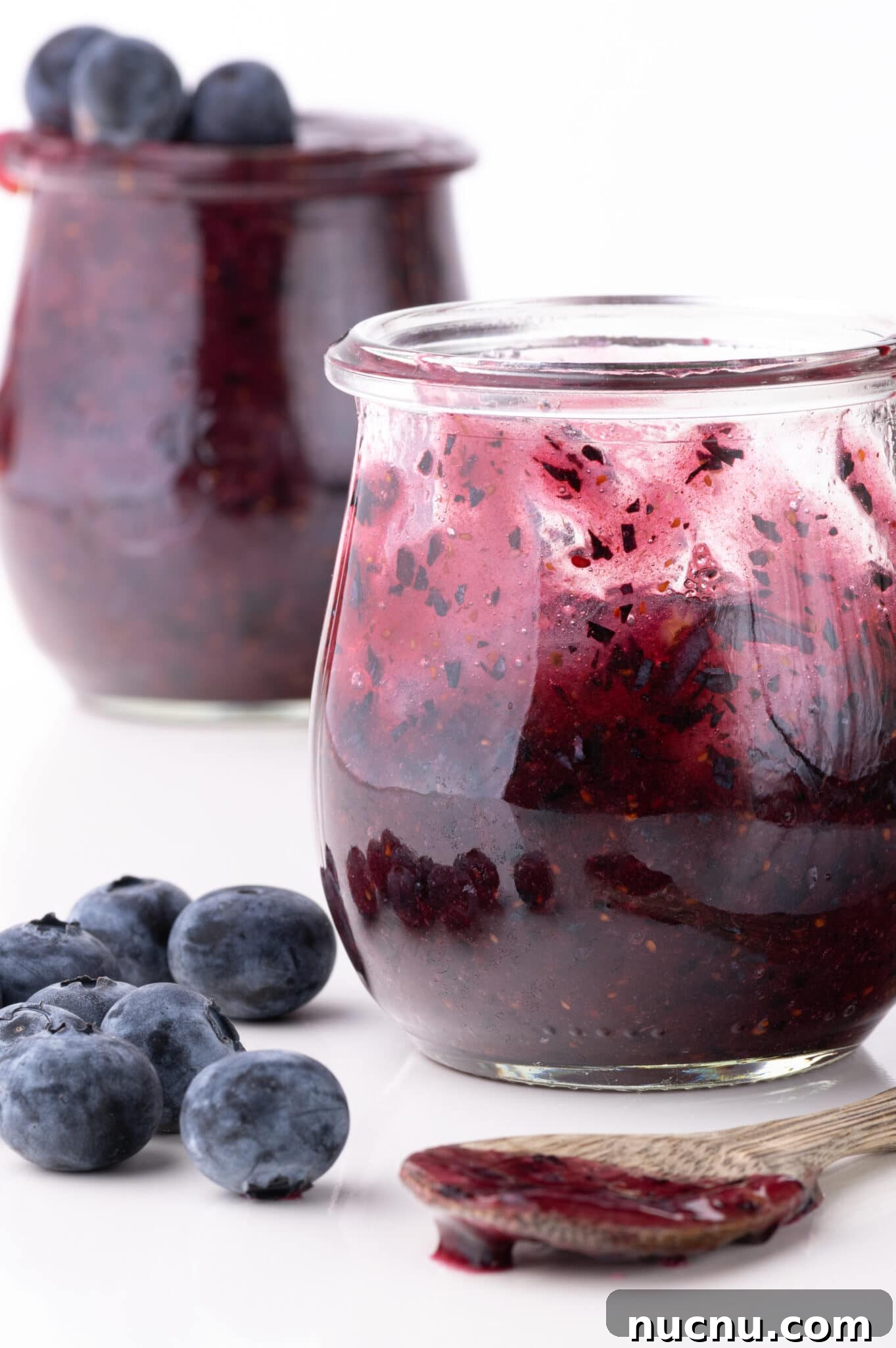 Keto Blueberry freezer jam in a small jar surrounded by fresh blueberries and a wooden spoon coated in fresh jam.