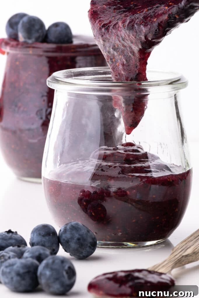 Small jars being filled with blueberry jam. The jars are against a bright white background with fresh plump blueberries in the foreground.