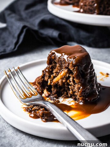 Close up of a mini cake on a white plate cut in half, revealing a moist crumb with bits of apples inside. The cake is covered in a drizzle of salted caramel sauce. There are other cakes on a platter in the background with a navy blue napkin.