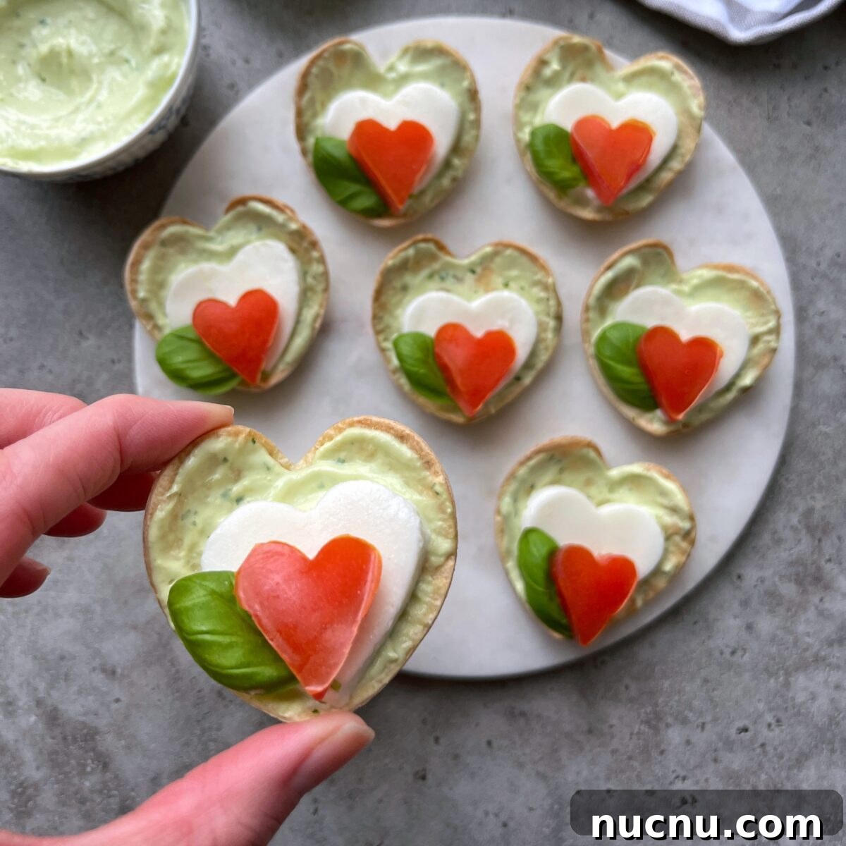 Caprese Bites: Fresh Tomato, Mozzarella & Basil Skewers 3 Heart-shaped Caprese Bites with green avocado spread, fresh basil, a mozzarella heart, and a tomato heart on top.