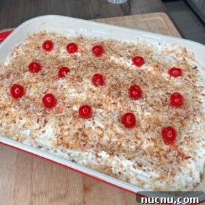 Pina Colada Cake Recipe with Coconut Rum Frosting 5 A rectangular dish filled with a dessert topped with toasted shredded coconut, whipped cream, and evenly spaced red maraschino cherries. The dish is on a wooden cutting board.