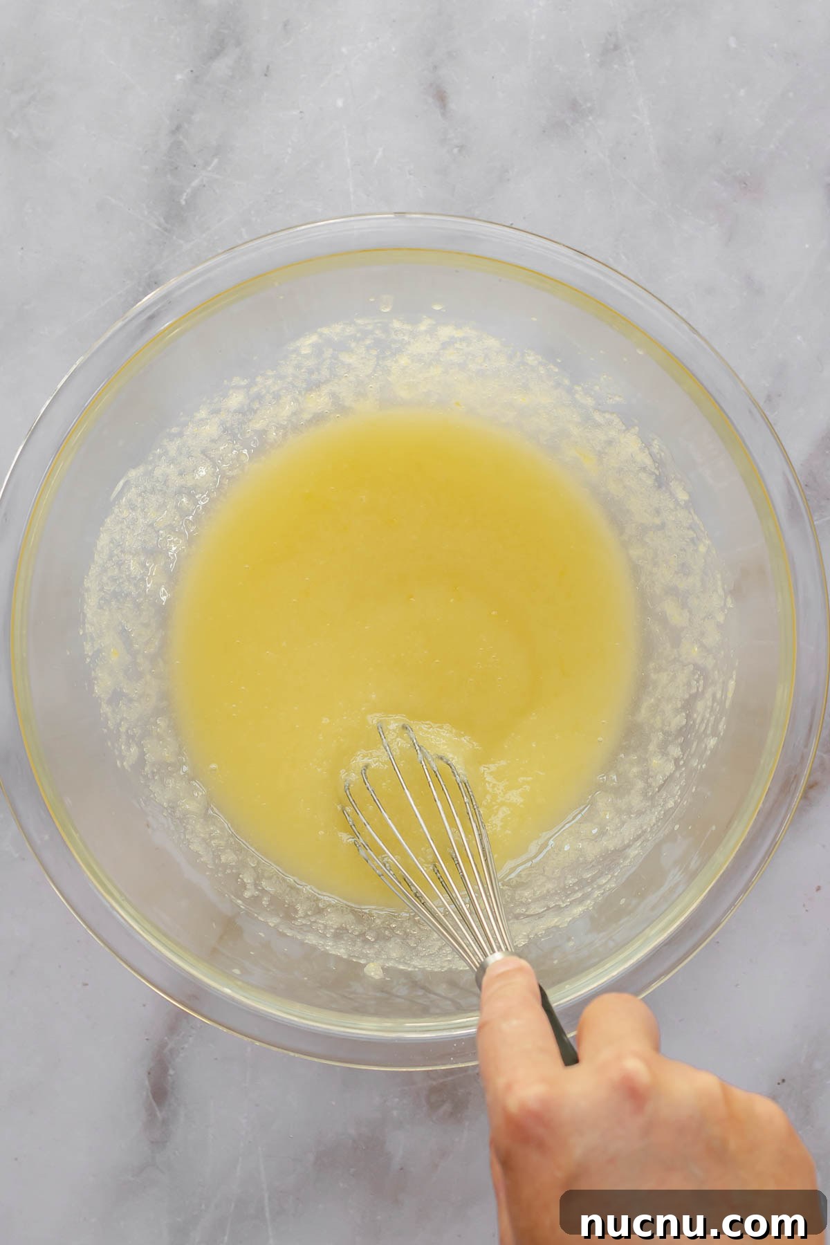 A hand whisks wet ingredients in a large glass bowl.