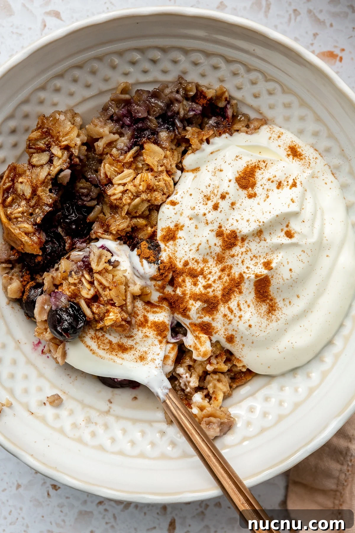 A serving of high protein oatmeal bake has been placed into a white serving bowl. A dollop of yogurt is added on top. A copper spoon is placed to the side.