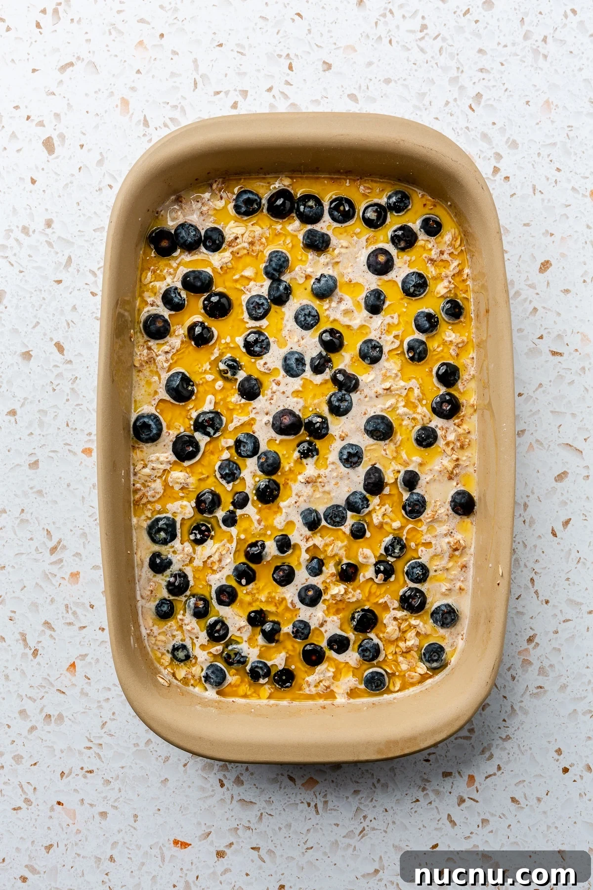 The liquid ingredients have been added over the top of the dry in the beige-colored baking dish. It has been topped with blueberries.