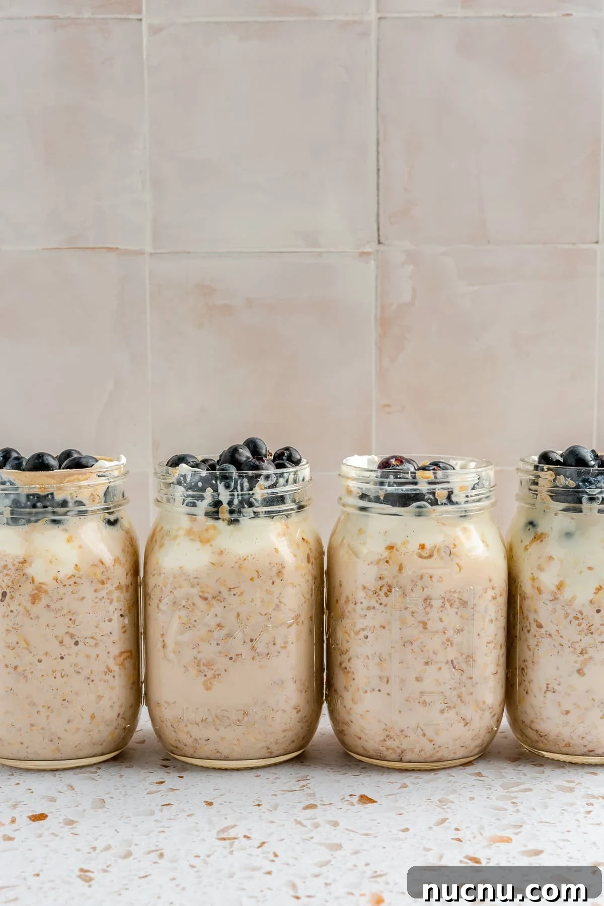 4 mason jars, full of high protein overnight oats, have been topped with blueberries. They sit arranged in a line on a white speckled countertop.