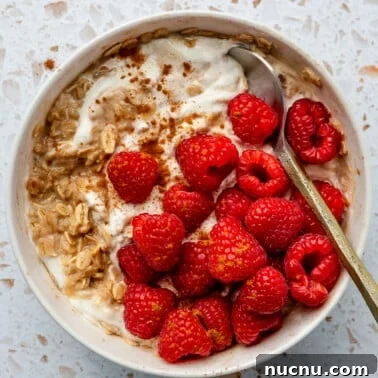A bowl of hot protein overnight oats sits in a white serving bowl. It has been topped with raspberries and a metal spoon.