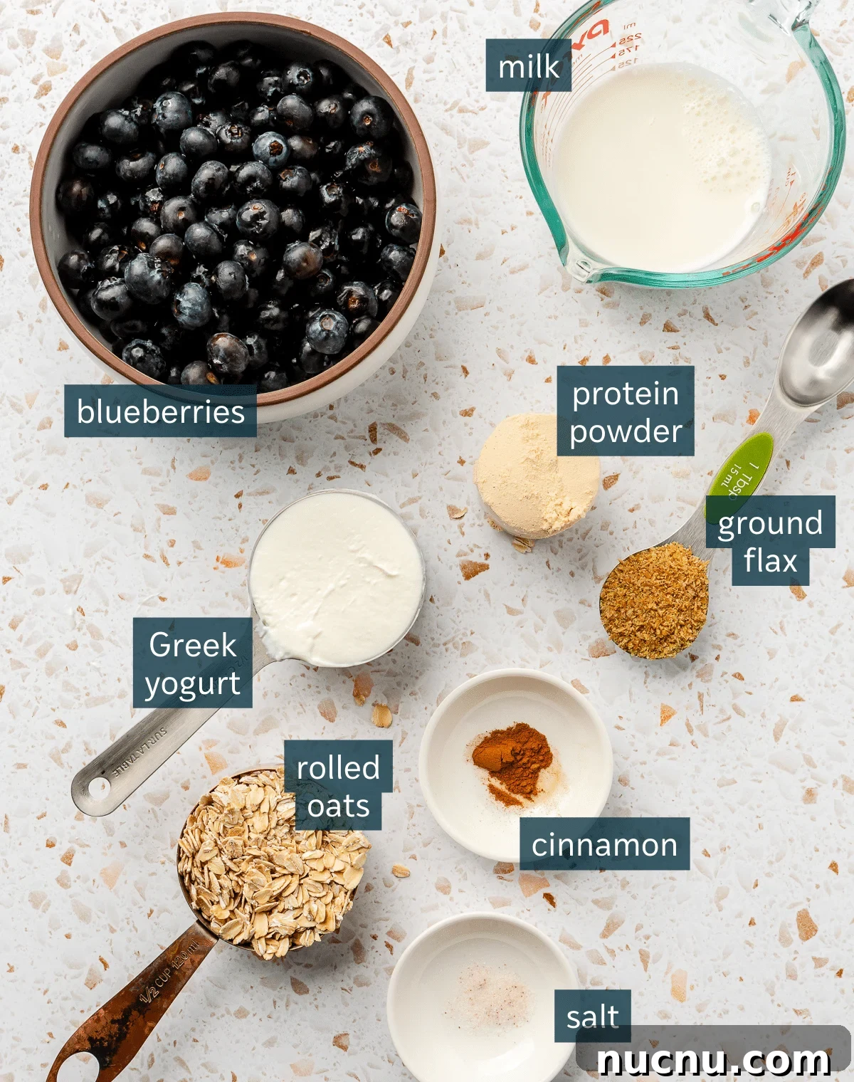 Ingredients for high protein overnight oats sit in a variety of bowls on a white speckled countertop.