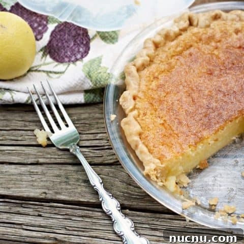 This is a photo of a Old Fashioned Lemon Chess Pie with a slice cut out a fork a lemon and a kitchen towel