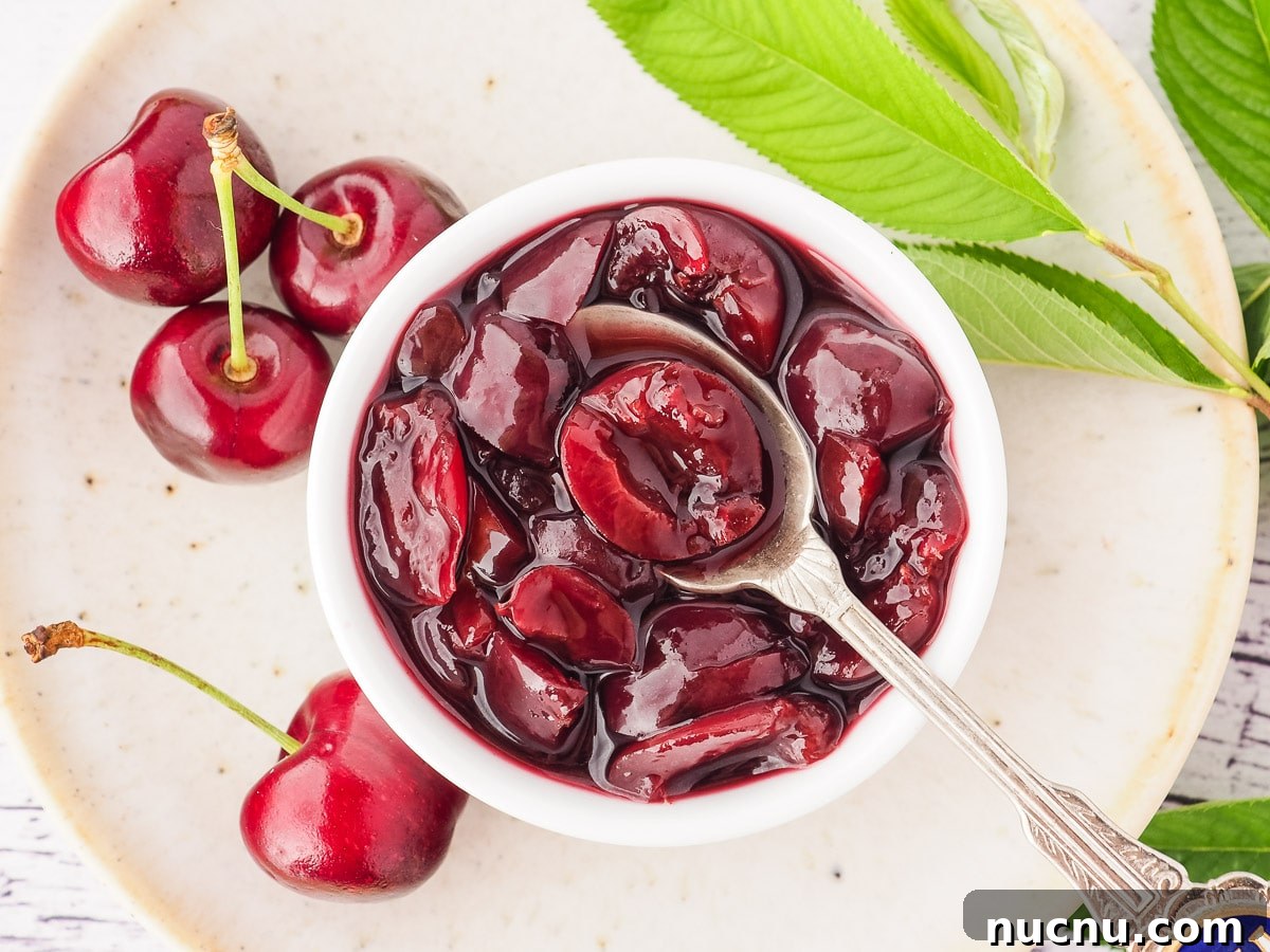 Homemade Cherry Compote Recipe for Desserts and Breakfast 3 Top down view of compote in a serving bowl with a spoon, on a plate with fresh cherries and cherry leaves.