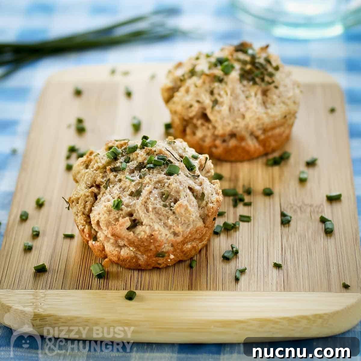 Side shot of two biscuits garnished with fresh chives on a wooden cutting board on a blue plaid cloth.