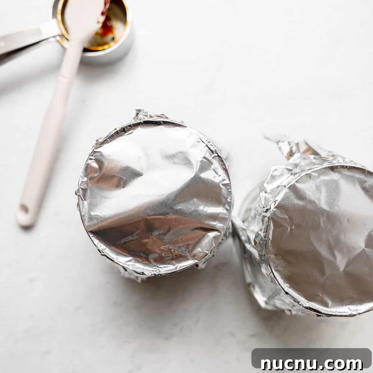 Authentic Boston Brown Bread Recipe: Steamed New England Classic 11 Two cans covered with aluminum foil on a light surface, next to a measuring spoon and a spatula.