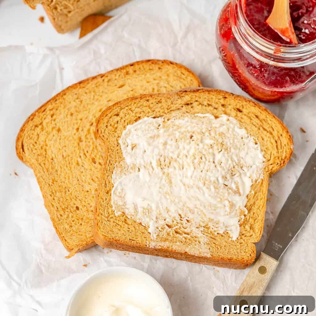 Authentic Boston Brown Bread Recipe: Steamed New England Classic 15 Two slices of bread on parchment paper, one spread with butter. A jar of red jam, a knife, and a small bowl of butter is nearby.