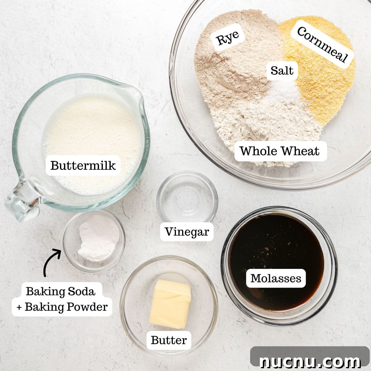 Authentic Boston Brown Bread Recipe: Steamed New England Classic 3 Top-down view of labeled ingredients for baking Boston Brown Bread: rye flour, cornmeal, whole wheat flour, salt, buttermilk, baking soda and powder, vinegar, butter, and molasses on a white surface.