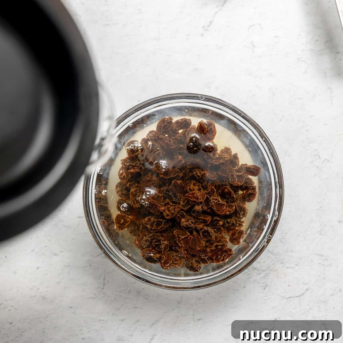 Authentic Boston Brown Bread Recipe: Steamed New England Classic 5 A glass bowl filled with raisins soaking in water on a light-colored surface.