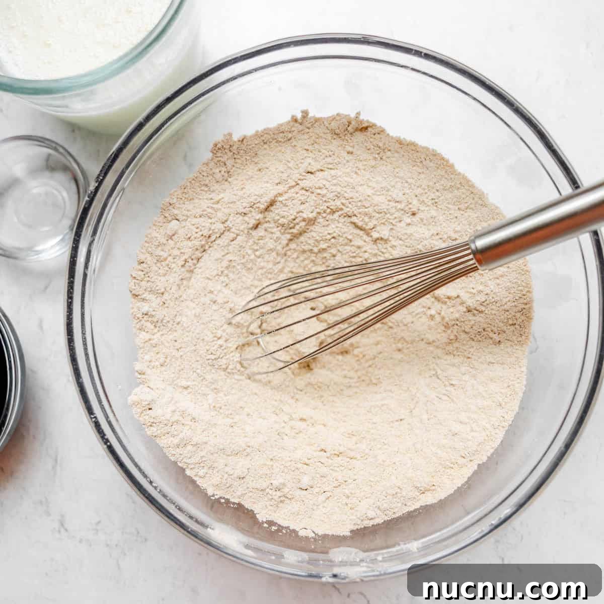 Authentic Boston Brown Bread Recipe: Steamed New England Classic 6 A metal whisk in a glass bowl of flour mixture on a white surface, with a glass measuring cup and small bowl nearby.
