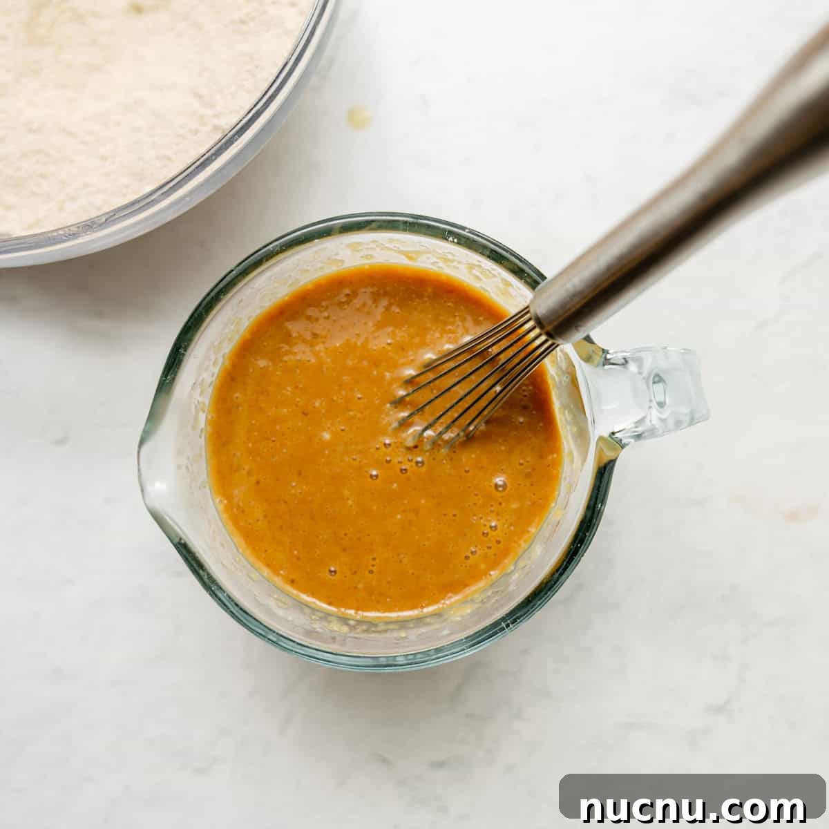 Authentic Boston Brown Bread Recipe: Steamed New England Classic 7 A metal whisk rests in a glass measuring cup filled with a smooth brown batter, with a bowl of dry ingredients visible in the background.