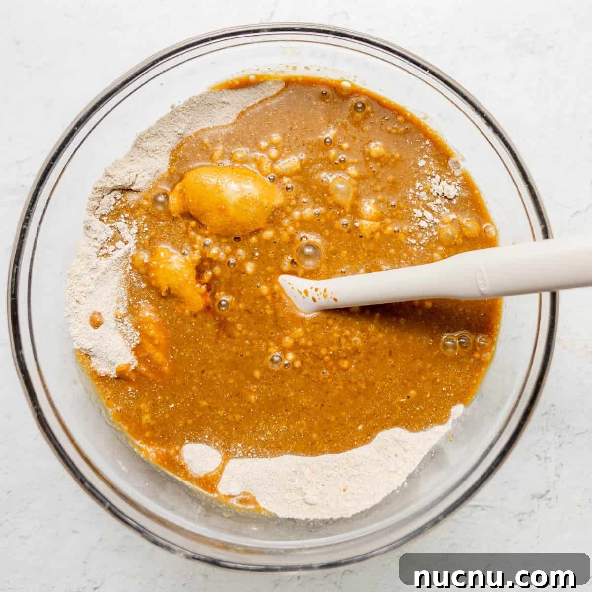 Authentic Boston Brown Bread Recipe: Steamed New England Classic 8 A glass bowl containing a mixture of flour and wet ingredients with a white spatula resting inside, ready for mixing.